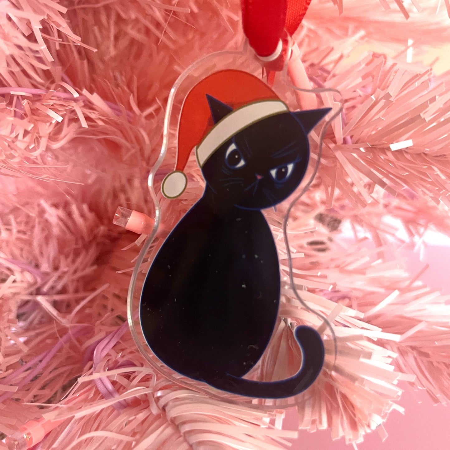 Grumpy black cat ornament with a Santa hat on pink tinsel background