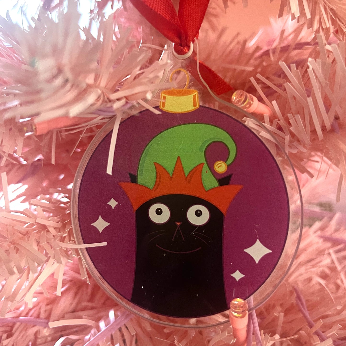 Decorative ornament with a black cat wearing a colorful hat on a pink tinsel background