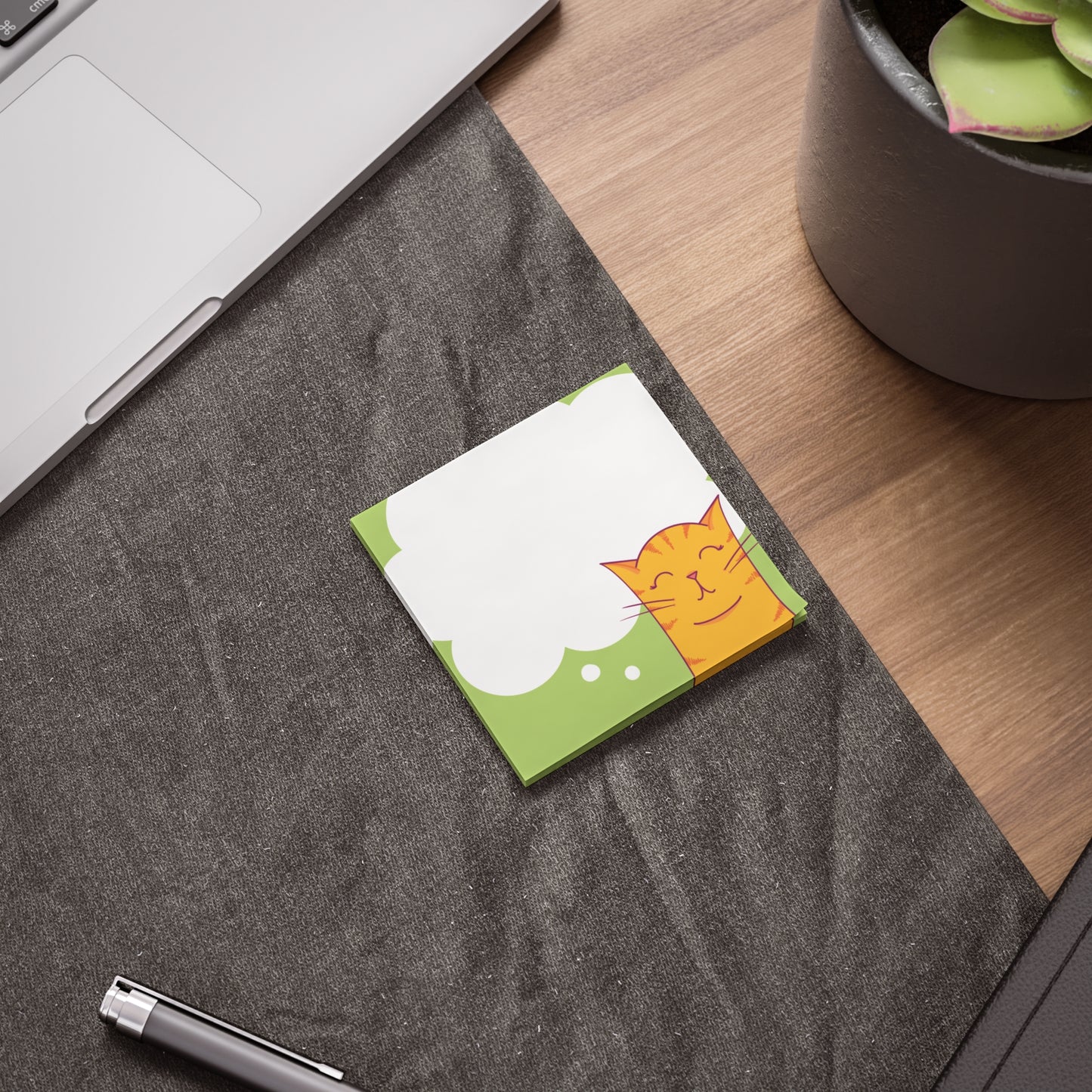 Small note pad with a cat illustration on a desk next to a laptop and plant